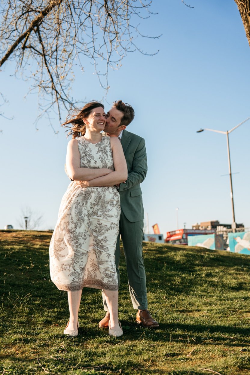 Tim hugs Hanna from behind and kisses her cheek on a grassy hill at golden hour